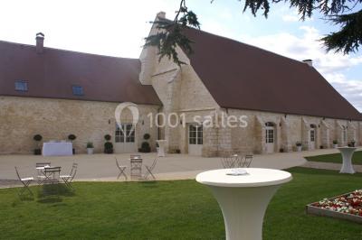 Bâtiment en pierre avec toit en pente, tables et chaises disposées sur une terrasse et une pelouse au premier plan.
