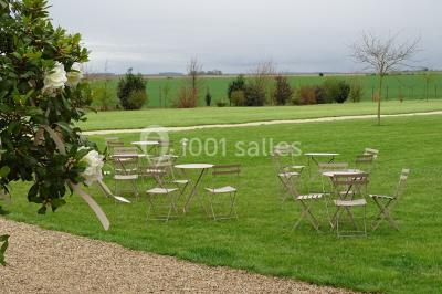 Bâtiment en pierre avec toit en pente, tables et chaises disposées sur une terrasse et une pelouse au premier plan.