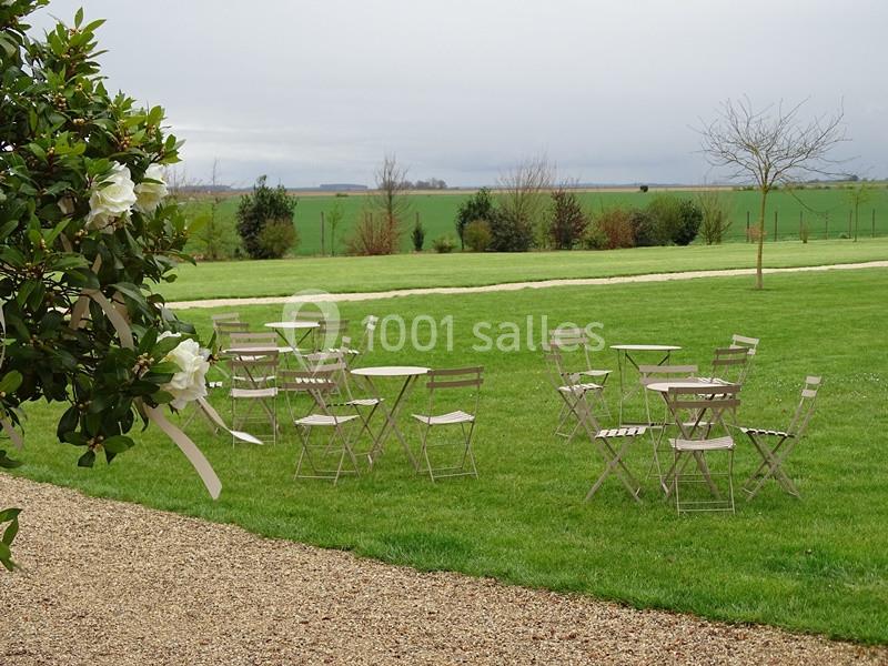 Chaises et tables en métal disposées sur une pelouse verte, avec un arbre fleuri au premier plan et un paysage rural en…