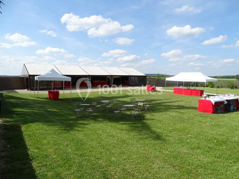 Tentes blanches et tables en métal disposées sur une pelouse verte sous un ciel bleu avec quelques nuages.