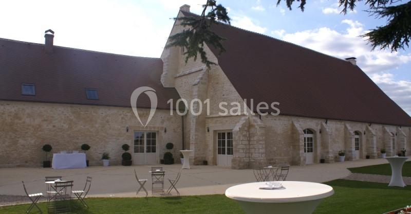 Bâtiment en pierre avec toit en pente, tables et chaises disposées sur une terrasse et une pelouse au premier plan.