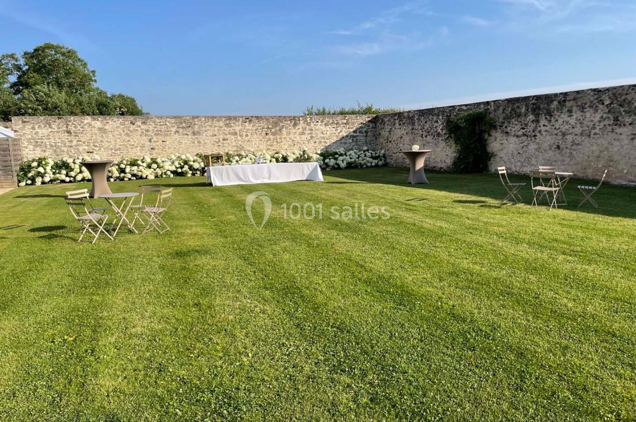 Jardin avec pelouse bien entretenue, tables et chaises dispersées, mur en pierre et ciel dégagé en arrière-plan.
