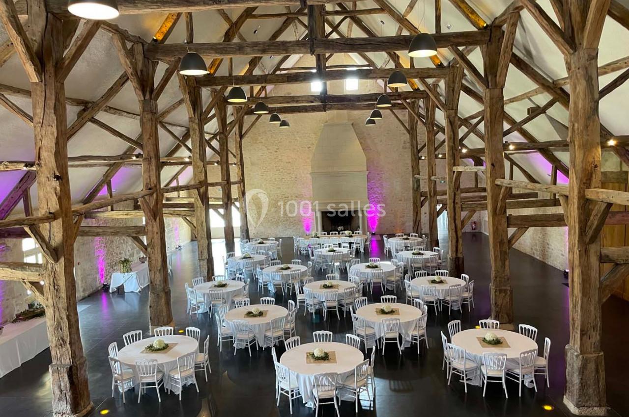 Grande salle de réception avec charpente en bois apparente, tables rondes dressées et éclairage tamisé.