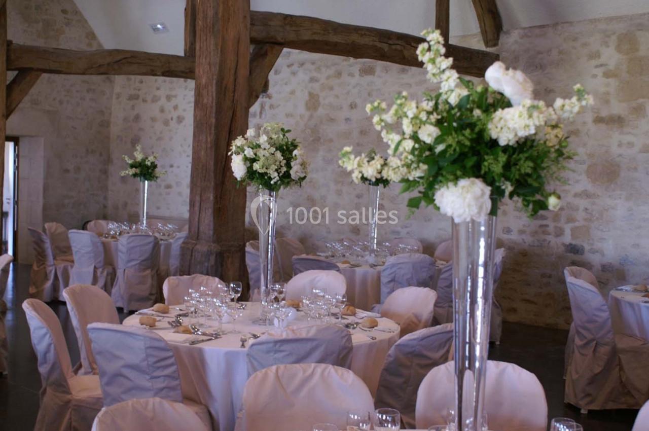Salle de réception avec tables rondes dressées, nappes blanches et centres de table fleuris dans des vases hauts.