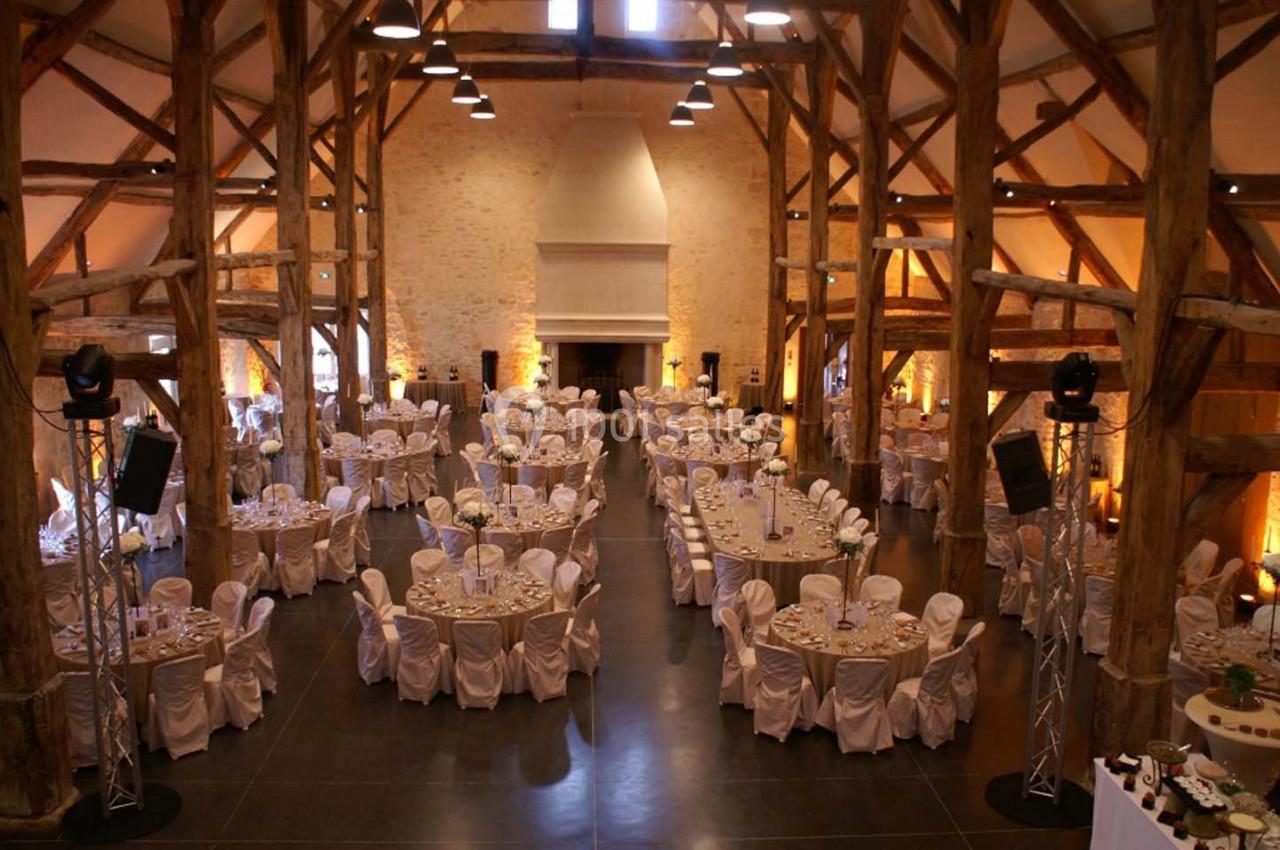 Salle de réception spacieuse avec charpente en bois, tables rondes dressées et chaises couvertes de housses blanches.