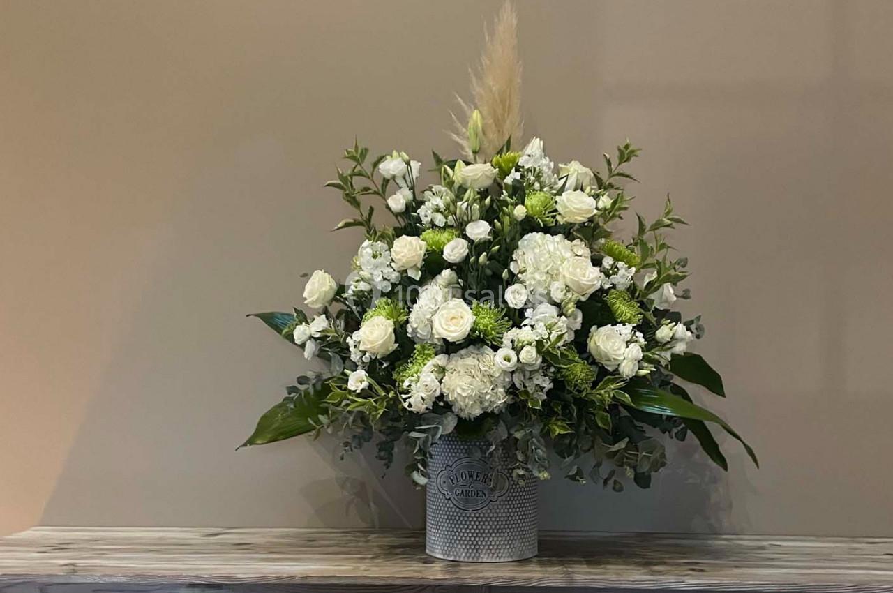 Bouquet de fleurs blanches et vertes dans un vase métallique, posé sur une table en bois devant un mur beige.