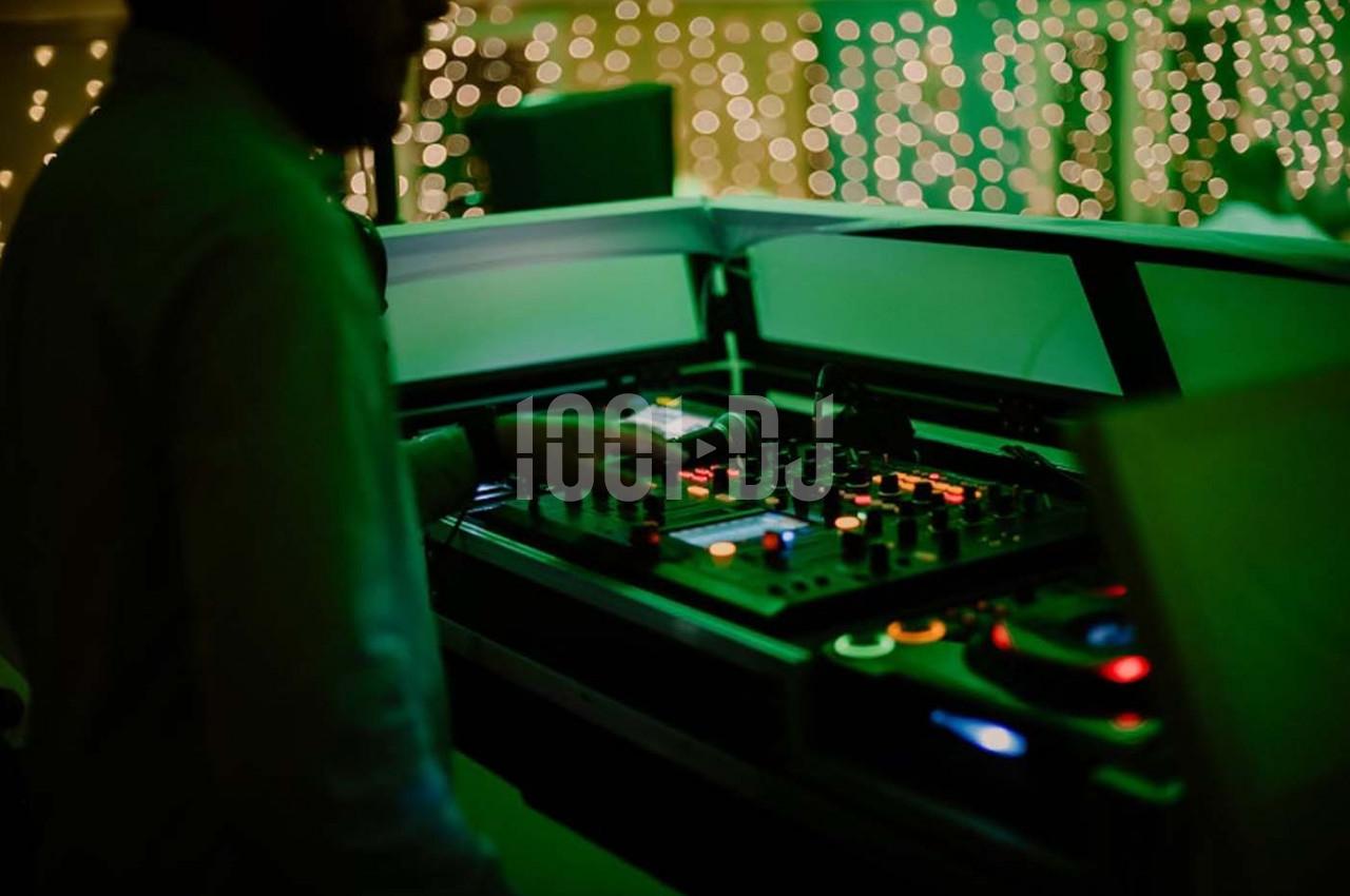 Un DJ manipule une table de mixage éclairée dans une ambiance tamisée avec des lumières floues en arrière-plan.