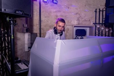 Un DJ mixe de la musique derrière une table éclairée, dans une salle en pierre avec des lumières colorées.