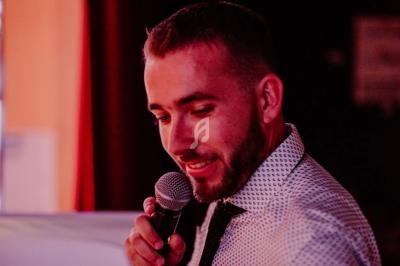Un homme en chemise blanche à motifs parle dans un micro dans un environnement faiblement éclairé.