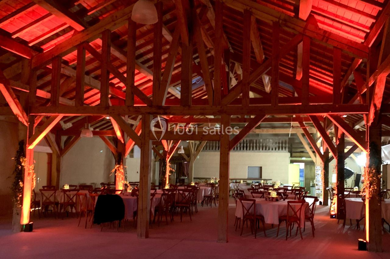 Salle de réception en bois avec tables rondes dressées et éclairage chaleureux rouge et orange.
