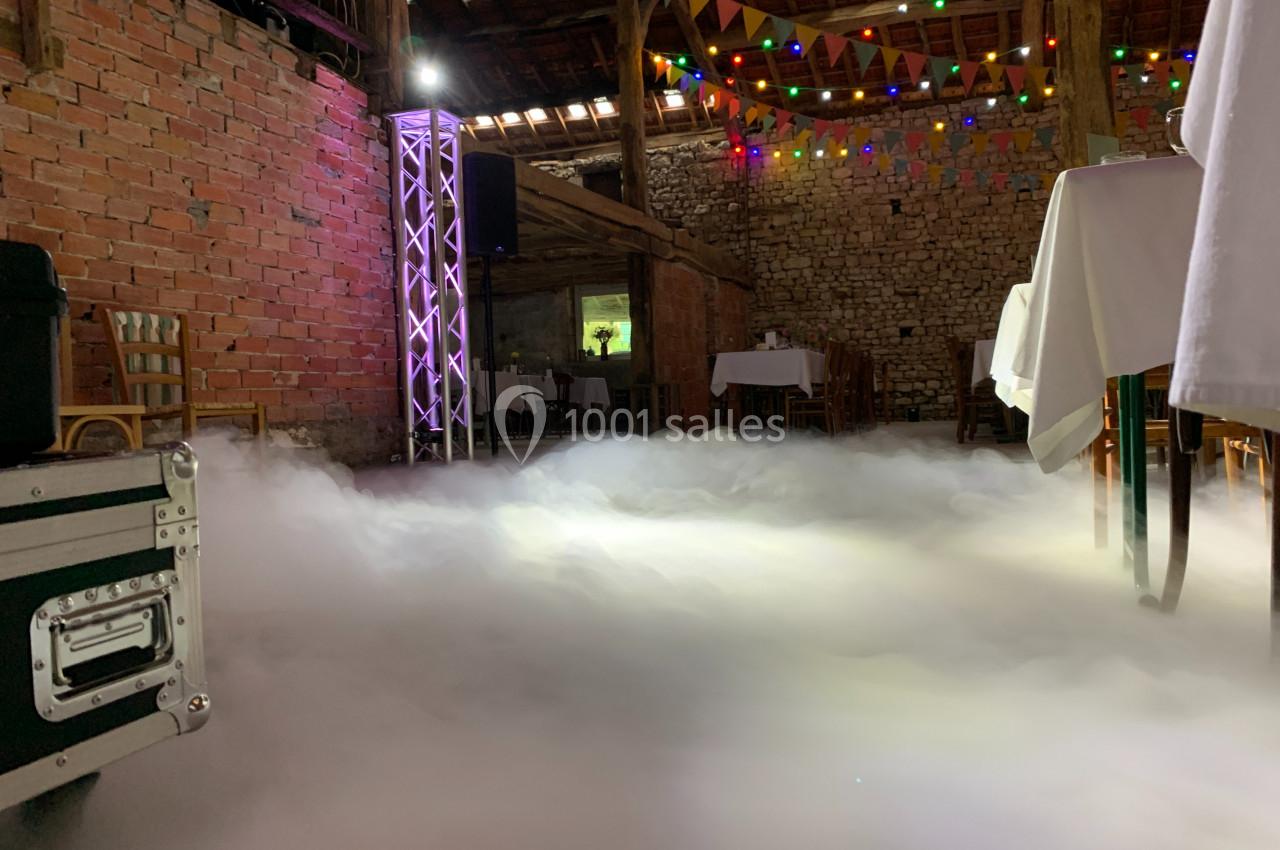 Salle en briques décorée pour un événement, avec tables dressées, guirlandes lumineuses et sol recouvert de fumée…