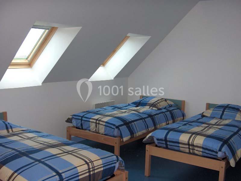 Trois lits simples avec des couvertures à carreaux bleus dans une chambre mansardée éclairée par des fenêtres de toit.