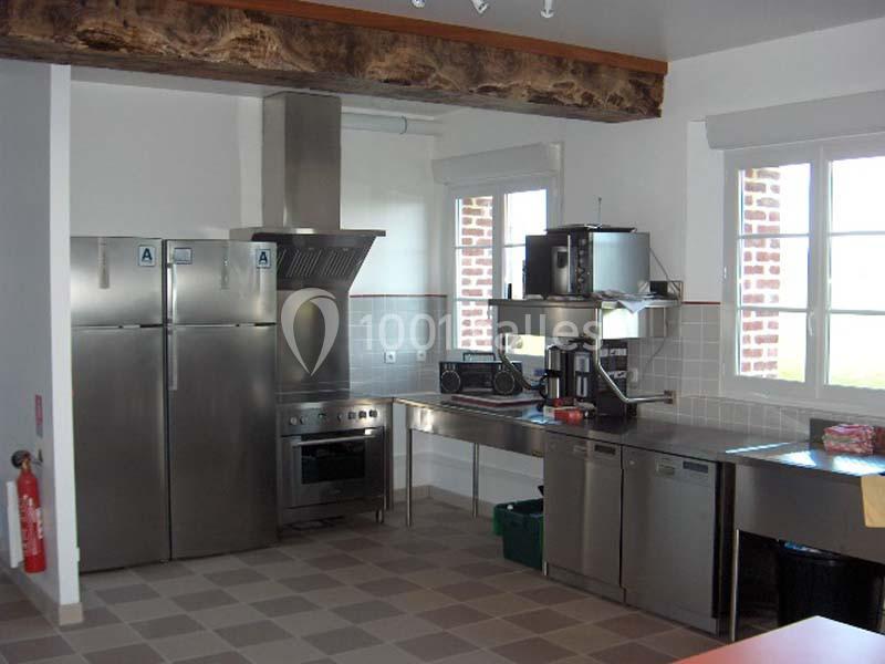 Cuisine équipée avec électroménagers en inox, plan de travail, hotte, réfrigérateurs et fenêtre laissant entrer la lumière.