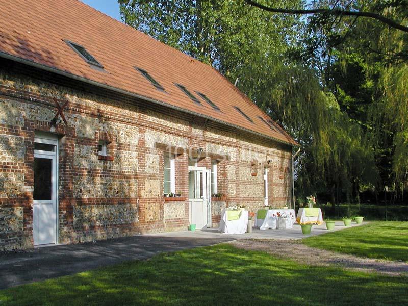 Bâtiment en briques avec toit en tuiles, terrasse aménagée avec tables et chaises, entouré de verdure.
