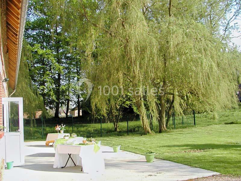 Terrasse ensoleillée avec tables dressées, bordée par un jardin verdoyant et des arbres, dont des saules pleureurs.