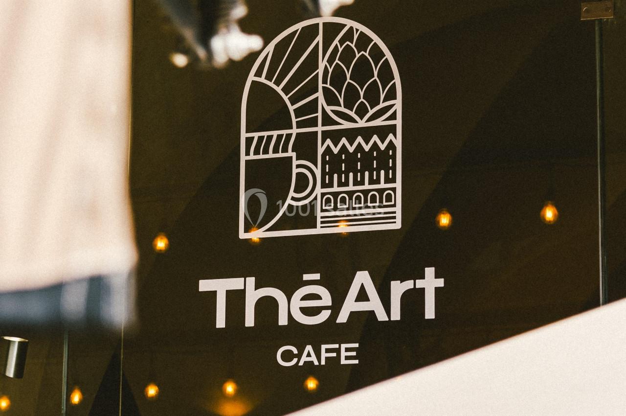 Logo ’The Art Café’ sur une vitre, entouré de lumières suspendues et d'éléments d'ombrage.