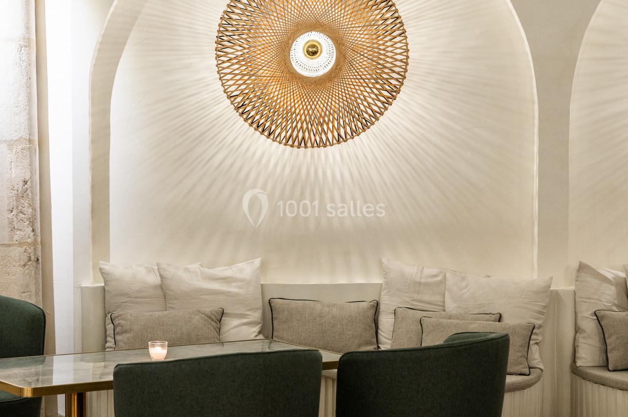 Espace intérieur avec fauteuils verts, table en verre, banquette beige et applique murale décorative en osier éclairée.