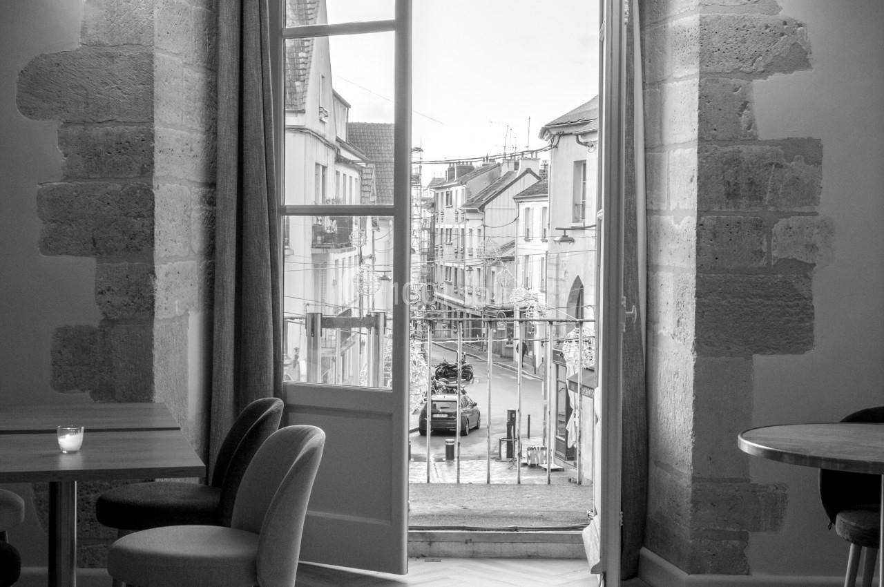 Salle lumineuse avec grandes fenêtres ouvertes sur une rue pavée, mobilier moderne et murs en pierre apparente.