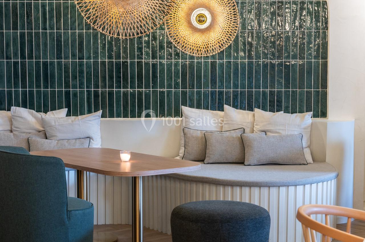 Coin salon avec table ronde, banquette, fauteuils et mur décoré de carreaux verts et luminaires dorés.