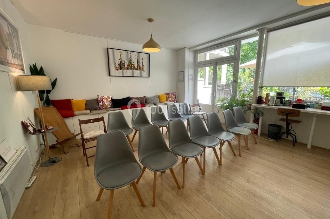 Salle lumineuse avec des chaises grises alignées, un canapé coloré en fond et une grande baie vitrée donnant sur un jardin.