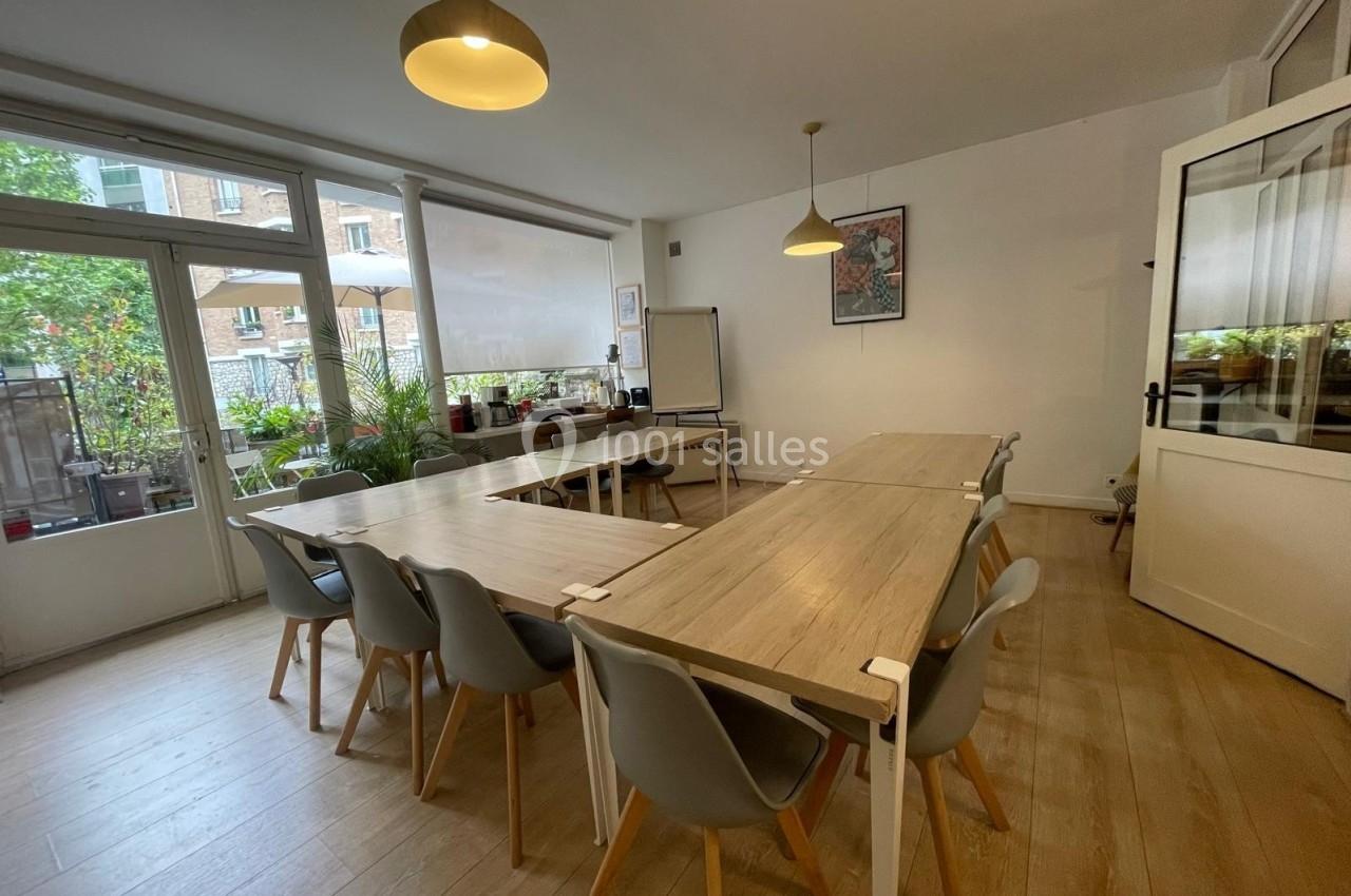 Salle lumineuse avec tables en bois, chaises grises, tableau blanc et vue sur une terrasse végétalisée.