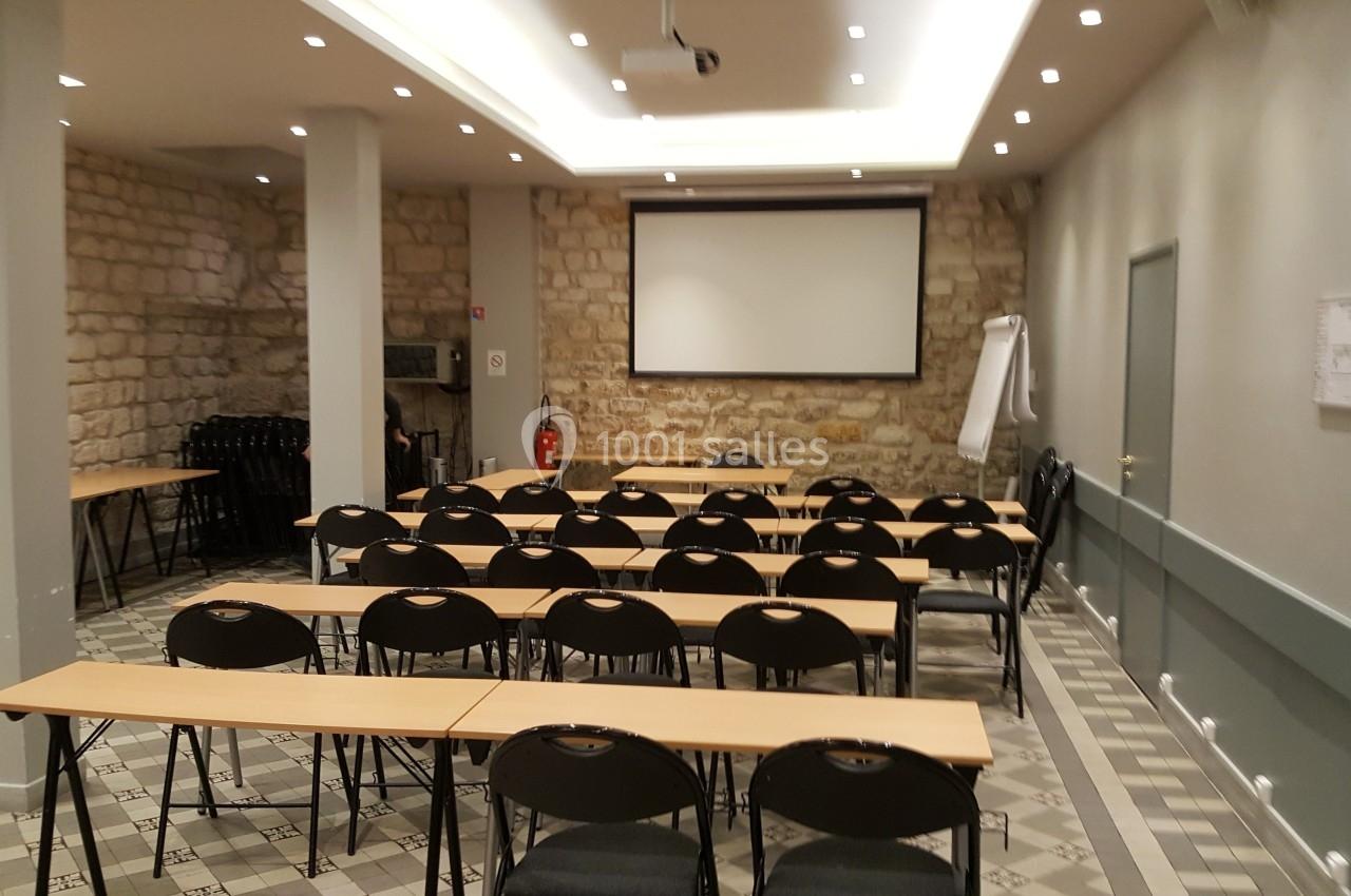Salle de réunion avec des rangées de tables et chaises, écran de projection et murs en pierre apparente.