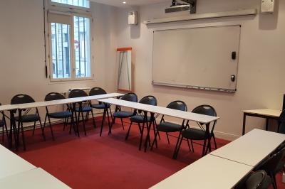 Salle de classe vide avec des tables blanches, des chaises bleues et un tableau de conférence au fond.