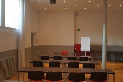 Salle de classe vide avec des tables blanches, des chaises bleues et un tableau de conférence au fond.
