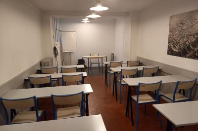 Salle de classe vide avec des tables blanches, des chaises bleues et un tableau de conférence au fond.