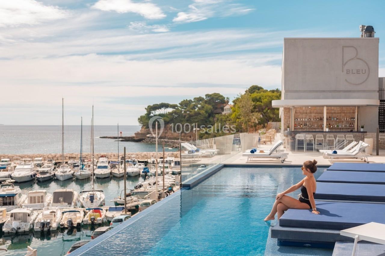 Une femme assise au bord d'une piscine surplombant un port de plaisance avec des bateaux et une vue sur la mer.