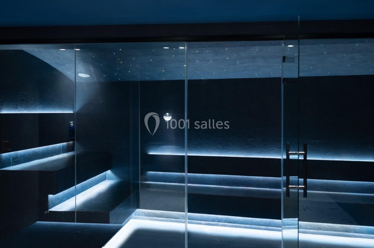 Salle de hammam moderne avec éclairage tamisé, murs sombres et bancs intégrés, vue à travers une porte vitrée.