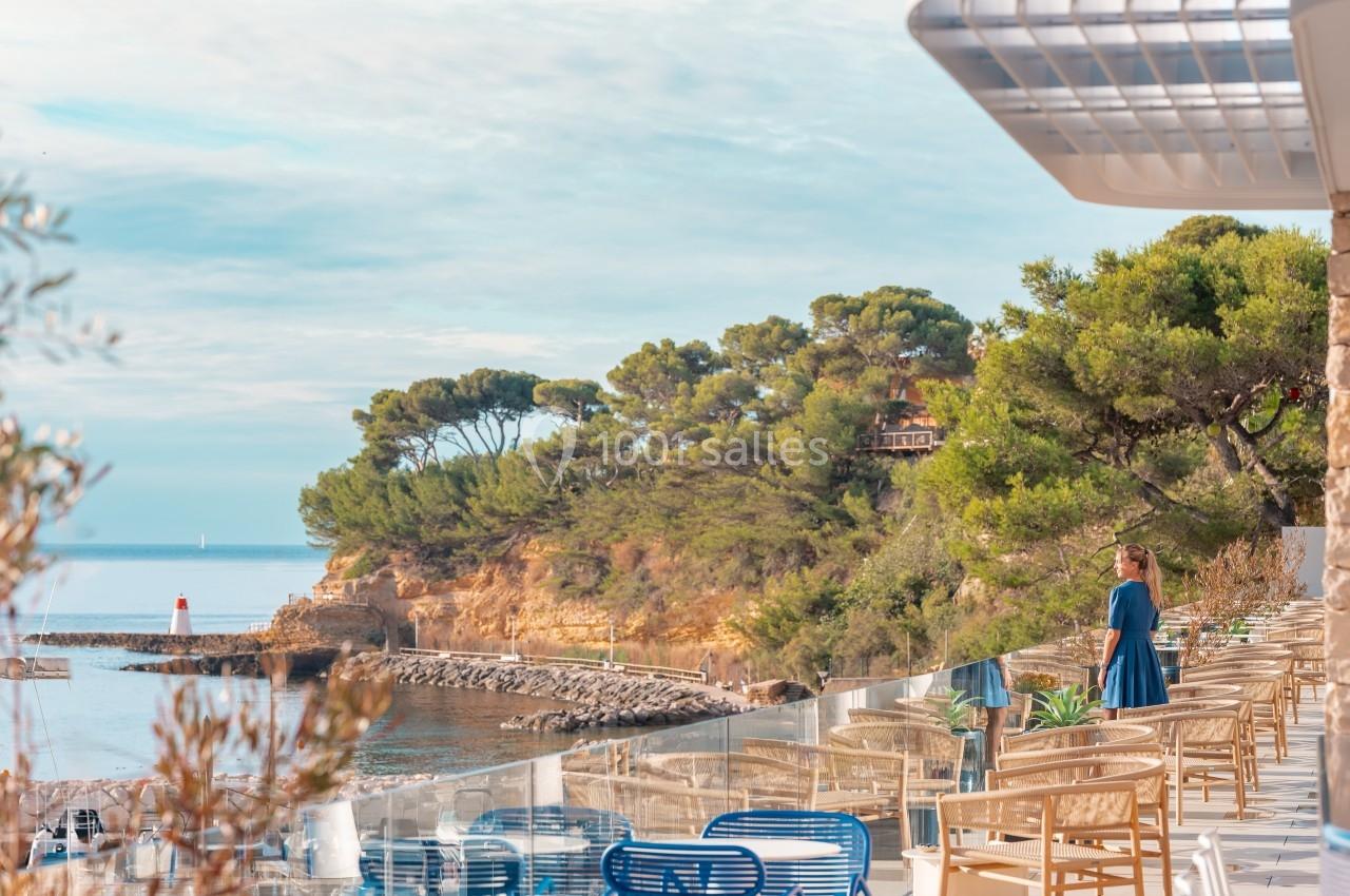 Vue d'une terrasse en bord de mer avec des tables et chaises, surplombant une côte boisée et un phare au loin.