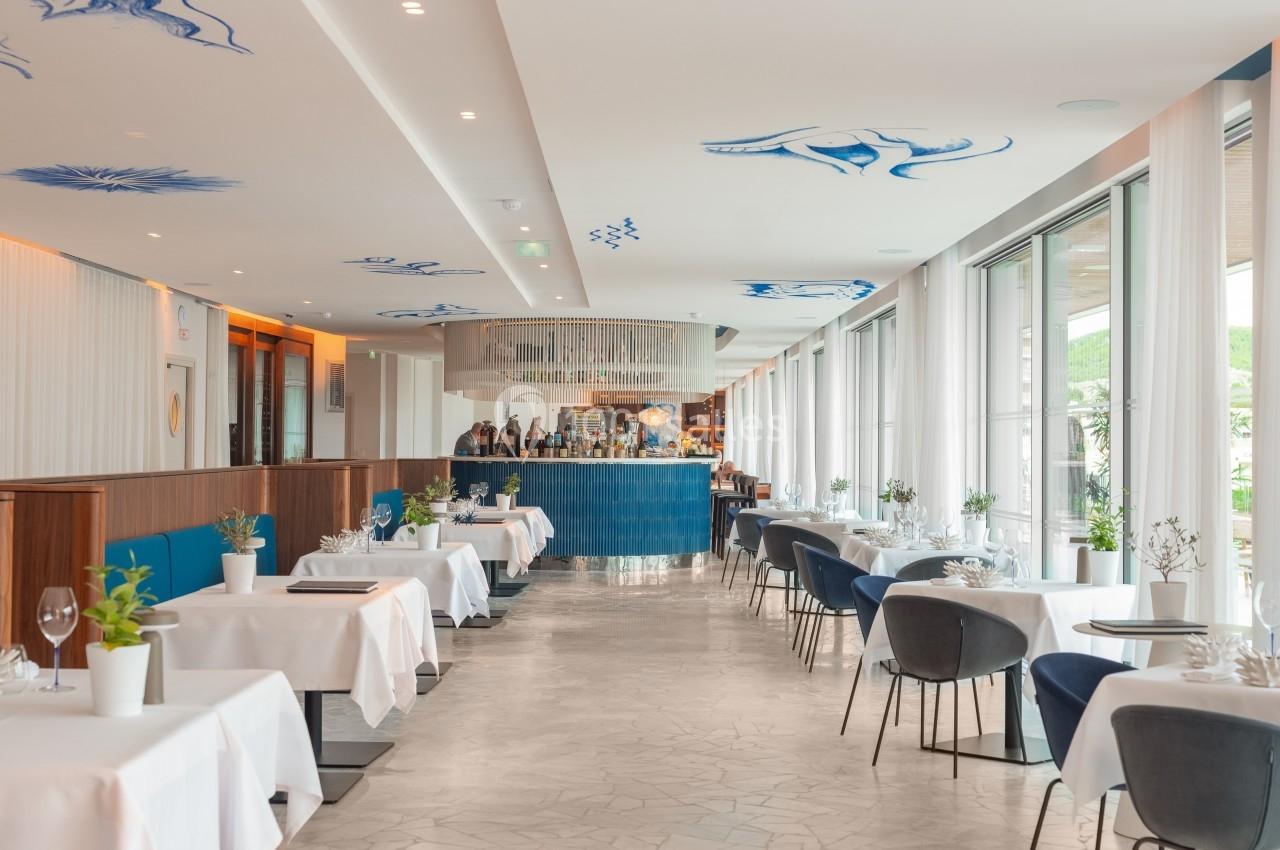 Salle de restaurant lumineuse avec tables dressées, chaises modernes et décoration bleue au plafond.