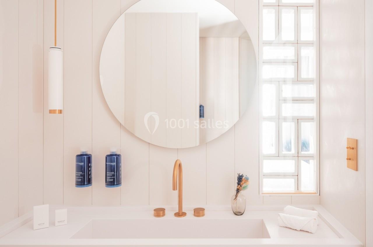Salle de bain minimaliste avec lavabo blanc, robinetterie dorée, miroir rond et flacons bleus fixés au mur.