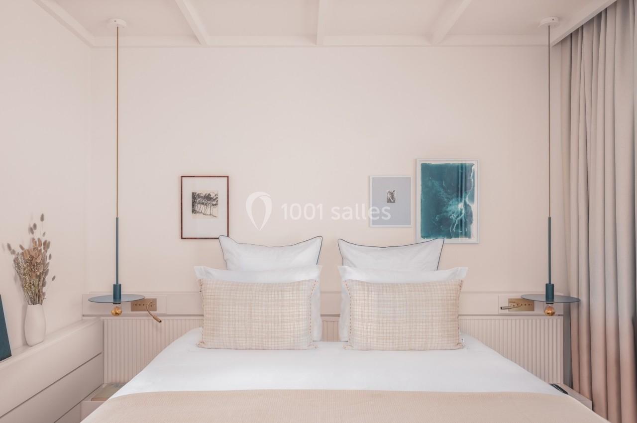 Lit double avec oreillers et coussins beiges, encadré par deux lampes suspendues et des tableaux sur un mur clair.