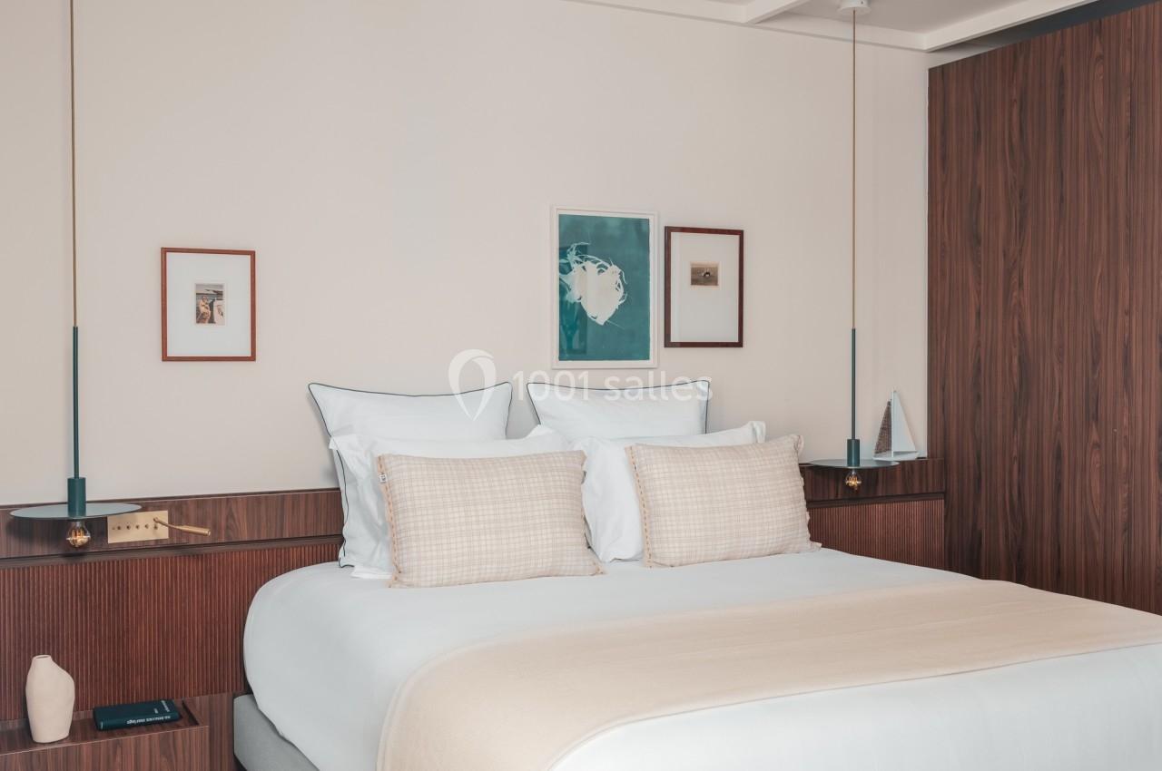 Lit double avec draps clairs, coussins beiges, tables de chevet en bois, décor minimaliste et cadres accrochés au mur.