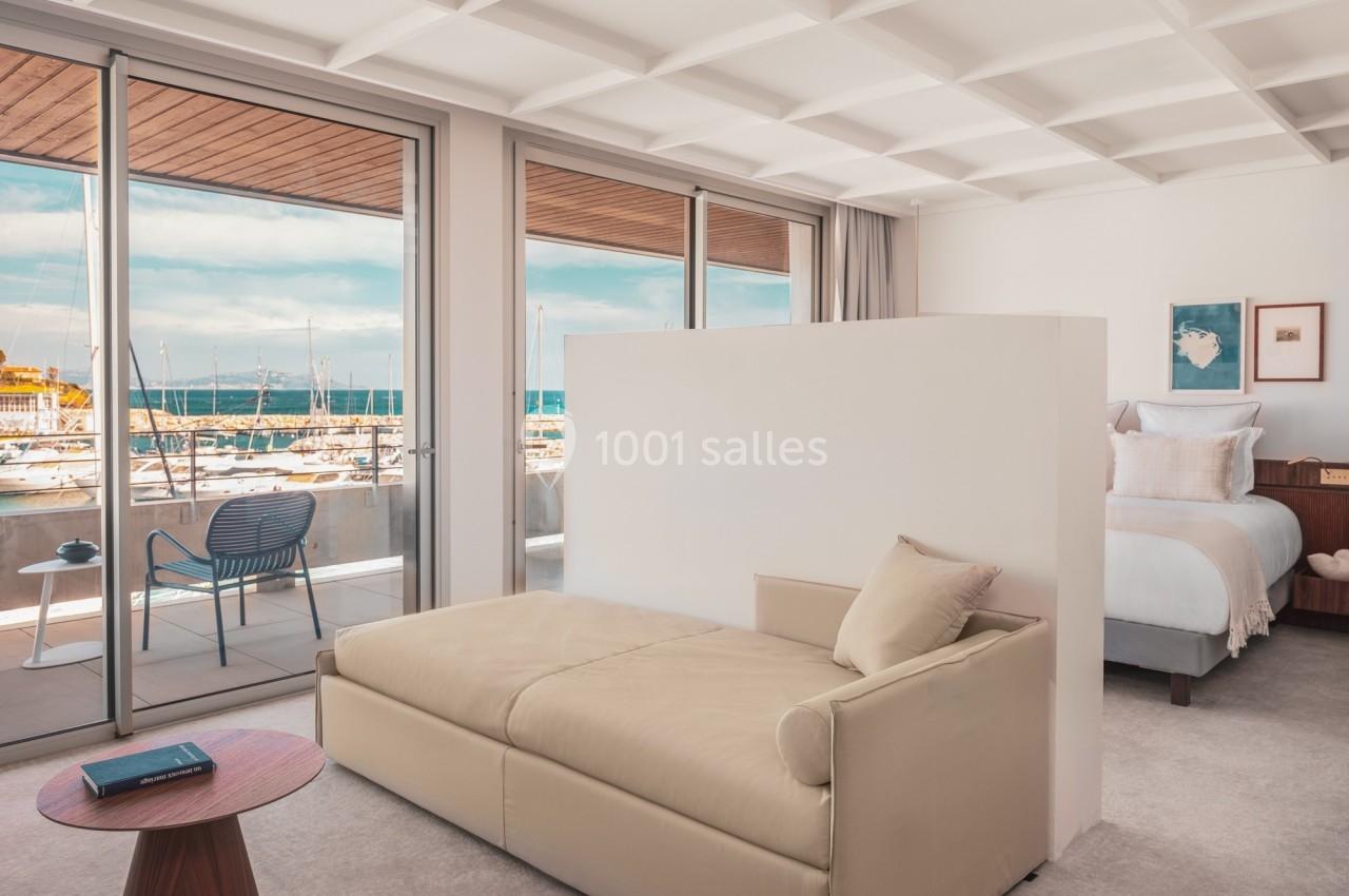 Vue d'une chambre lumineuse avec un canapé, un lit, et de grandes baies vitrées donnant sur un port de plaisance.