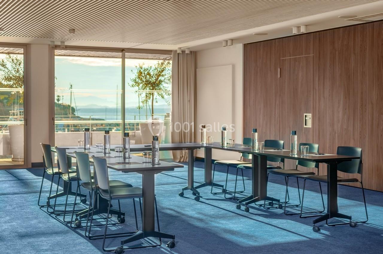 Salle de réunion lumineuse avec tables disposées en U, chaises modernes et vue sur un paysage extérieur ensoleillé.