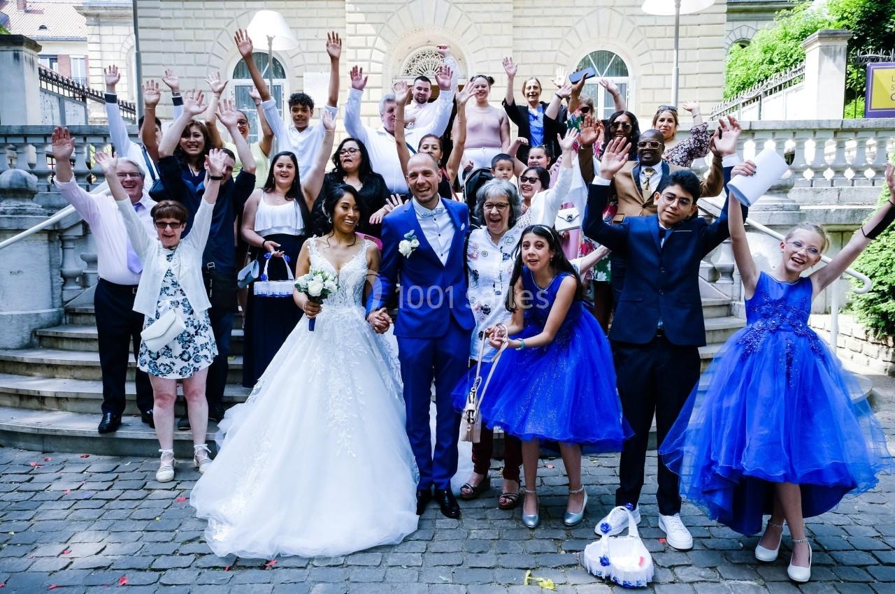 Un couple en tenue de mariage pose avec un groupe de personnes souriantes levant les bras devant un bâtiment.