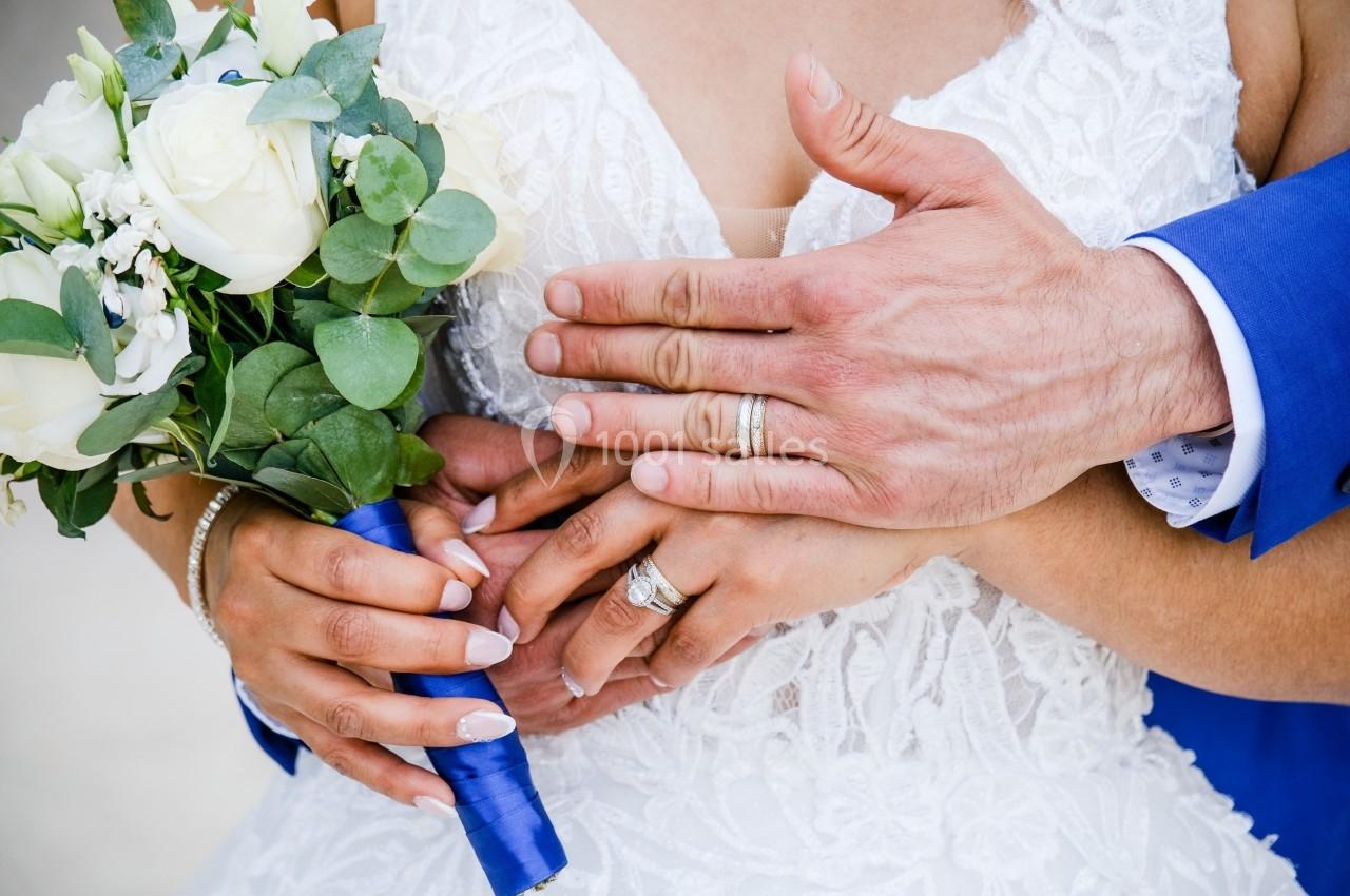 Mains d'un couple marié portant des alliances, tenant un bouquet de fleurs blanches et vertes avec un ruban bleu.