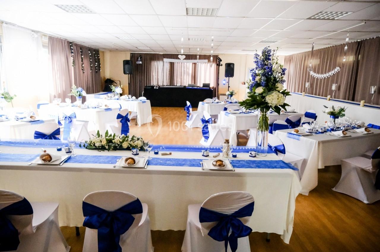Salle de réception décorée en blanc et bleu avec tables dressées, chaises ornées de nœuds et arrangements floraux.