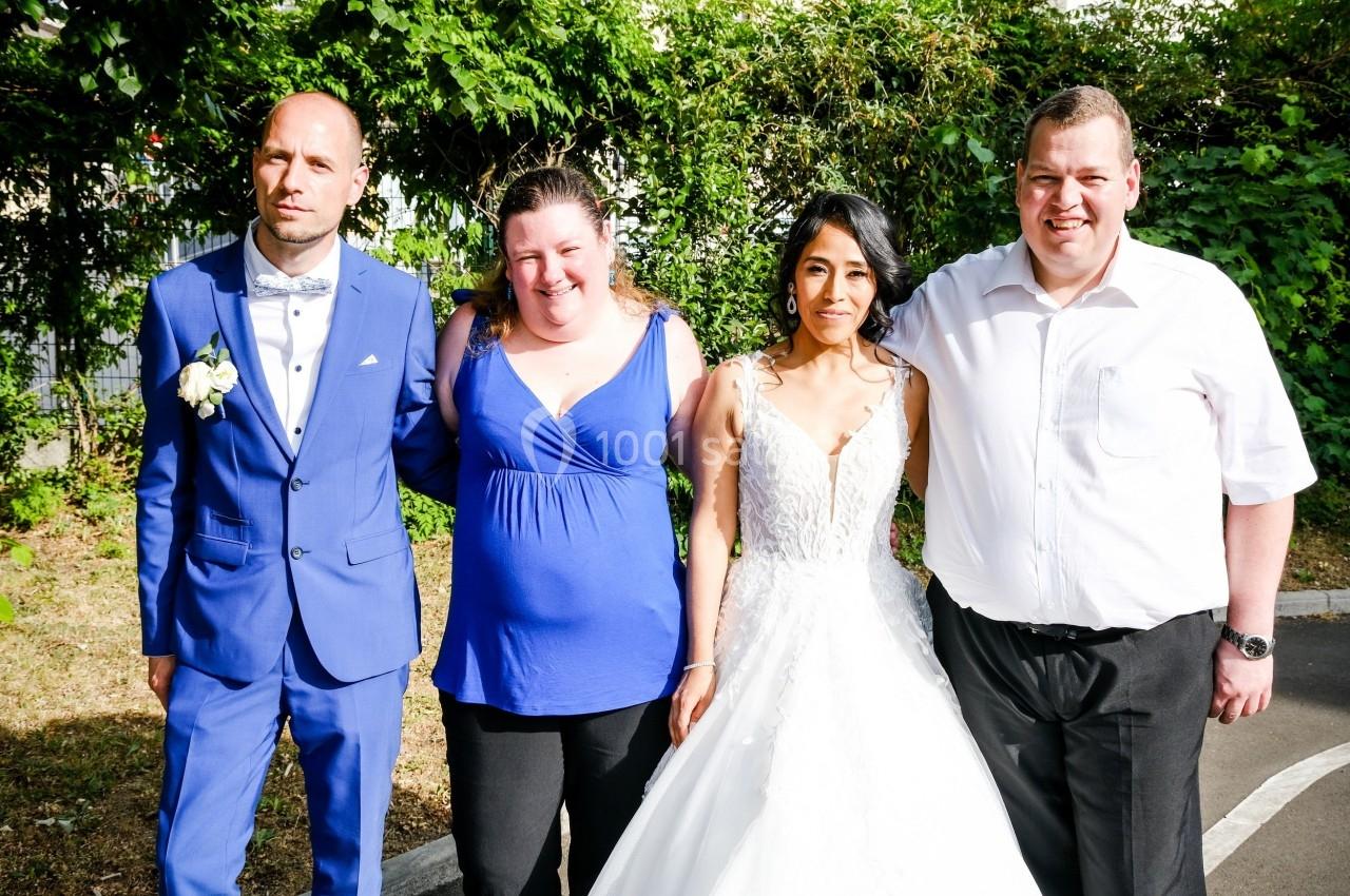 Quatre personnes posent debout en extérieur, dont une mariée en robe blanche et un homme en costume bleu.