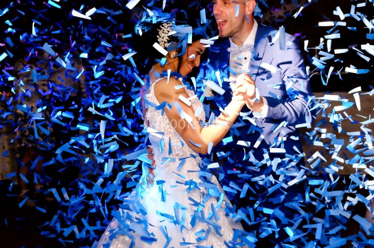 Un couple en tenue de mariage danse sous une pluie de confettis bleus dans une ambiance festive.