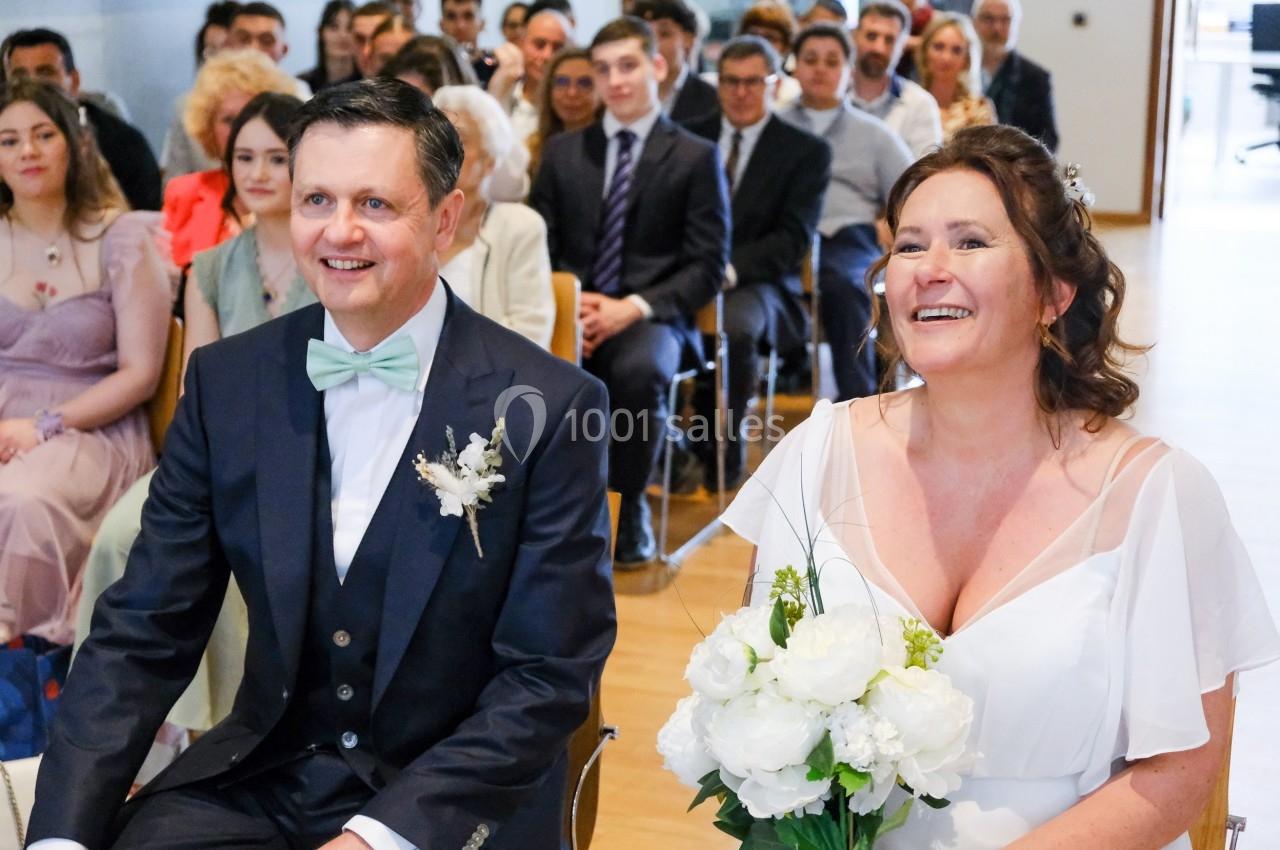 Un couple souriant assis lors d'une cérémonie de mariage, entouré d'invités attentifs en arrière-plan.