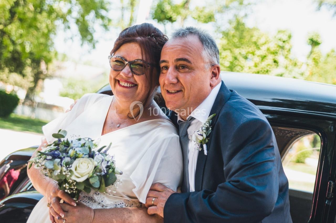 Un couple souriant en tenue de mariage pose devant une voiture ancienne dans un cadre extérieur ensoleillé.