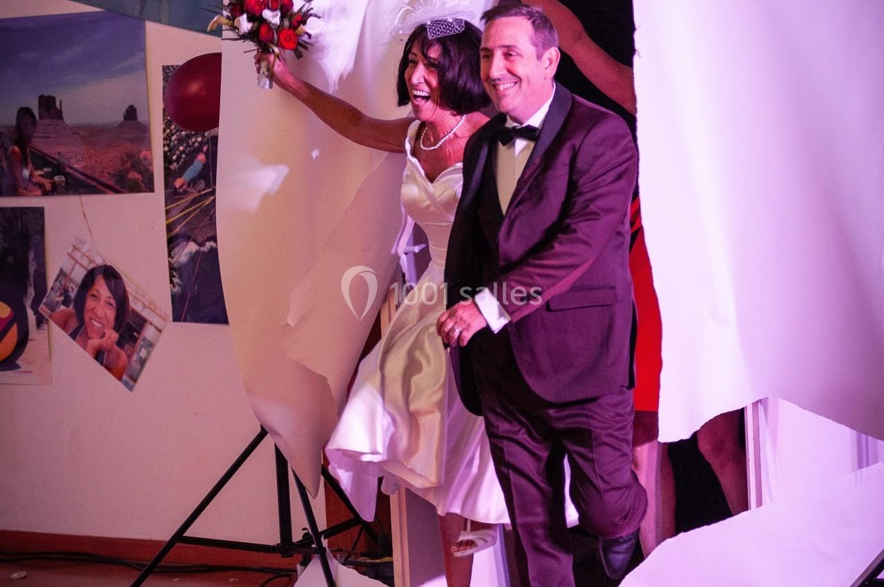 Un couple en tenue de mariage traverse une grande feuille de papier déchirée dans une salle décorée de photos.