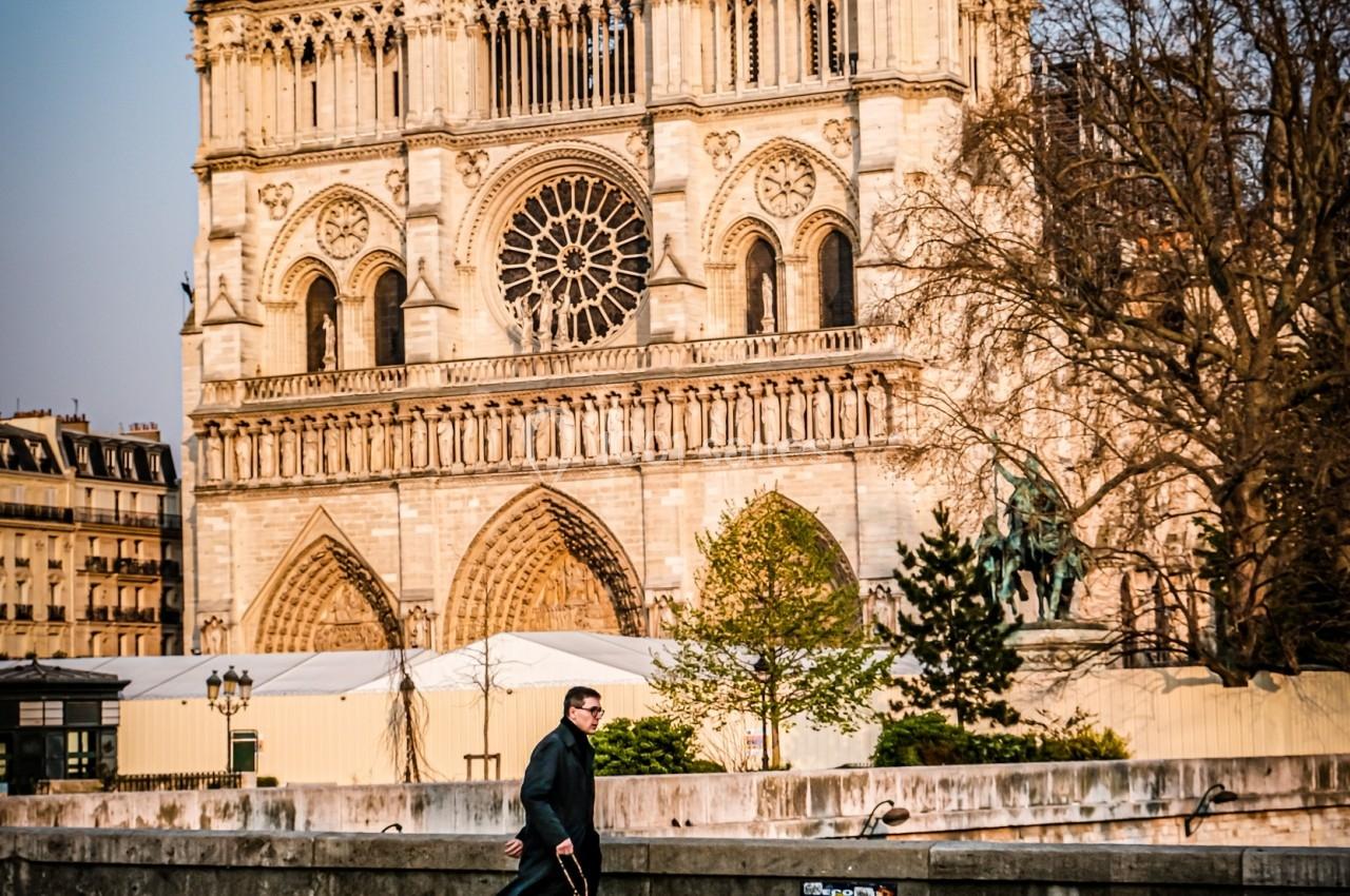 Un homme en manteau noir marche devant la cathédrale Notre-Dame de Paris par une journée ensoleillée.