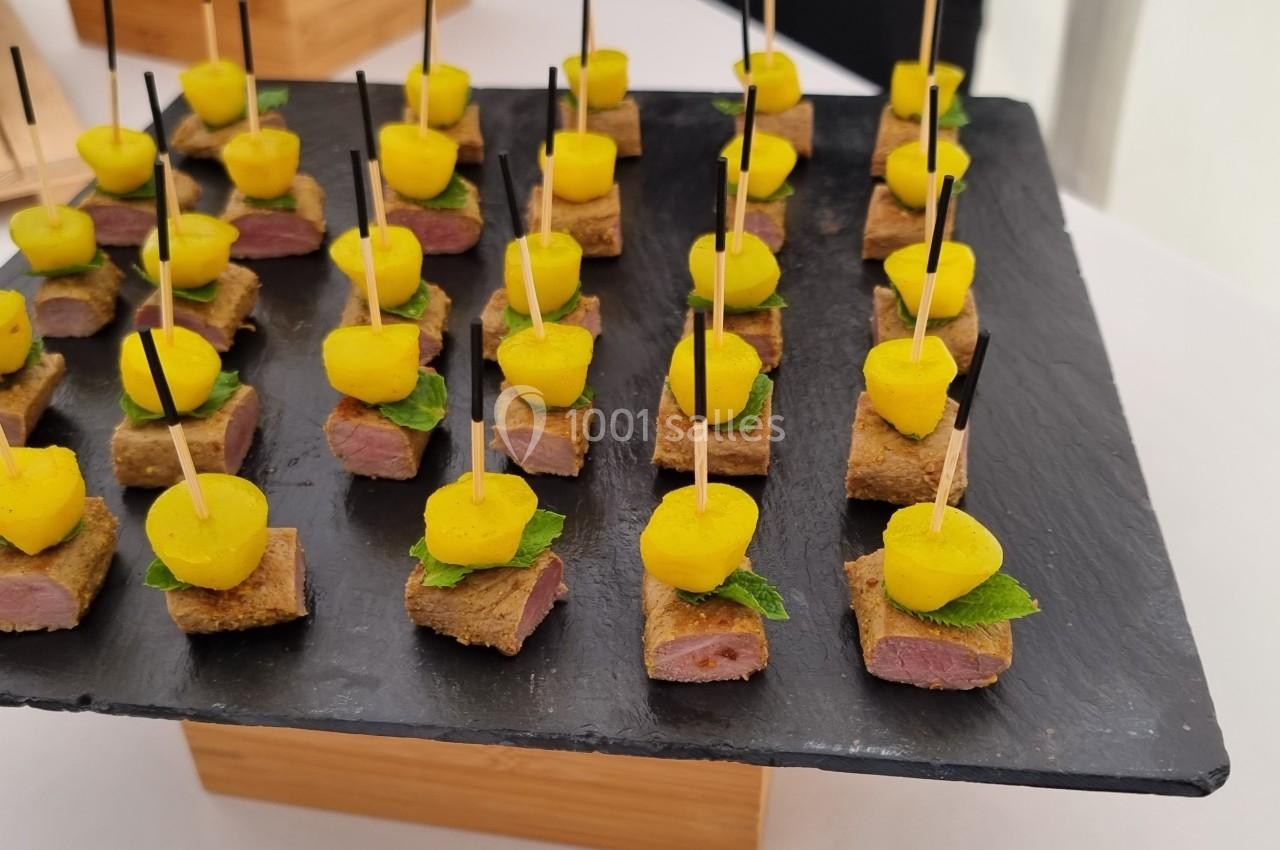 Bouchées apéritives sur ardoise, composées de viande, morceaux de mangue et feuilles de menthe, piquées sur des brochettes.