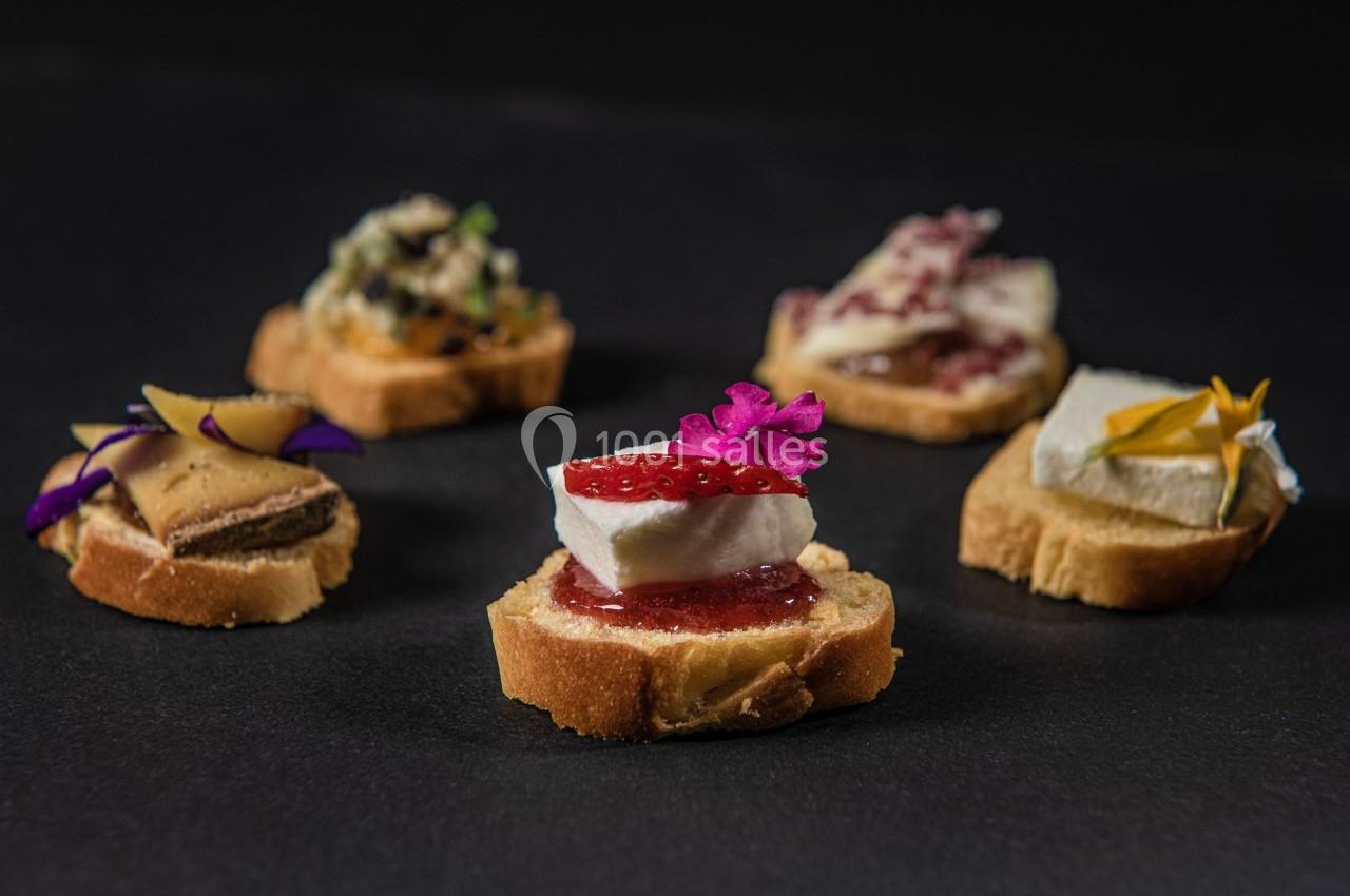 Assortiment de canapés garnis de fromage, confiture, fleurs comestibles et autres ingrédients variés sur fond sombre.