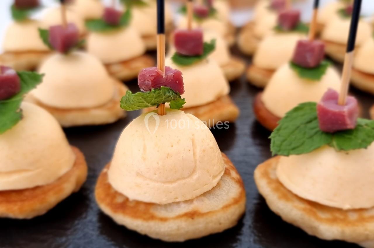 Petits canapés sur blinis garnis de mousse, feuilles de menthe et cubes de viande, présentés sur un plateau noir.
